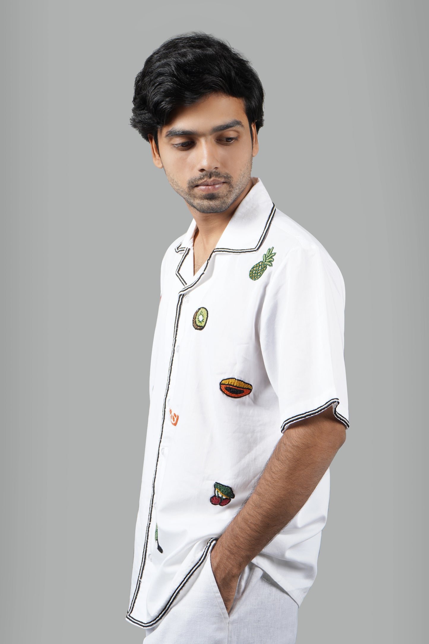 "White Cuban collar shirt featuring colorful hand-embroidered fruits like avocado, orange, kiwi, and pomegranate. Stylish and lightweight for a casual yet unique statement look."
