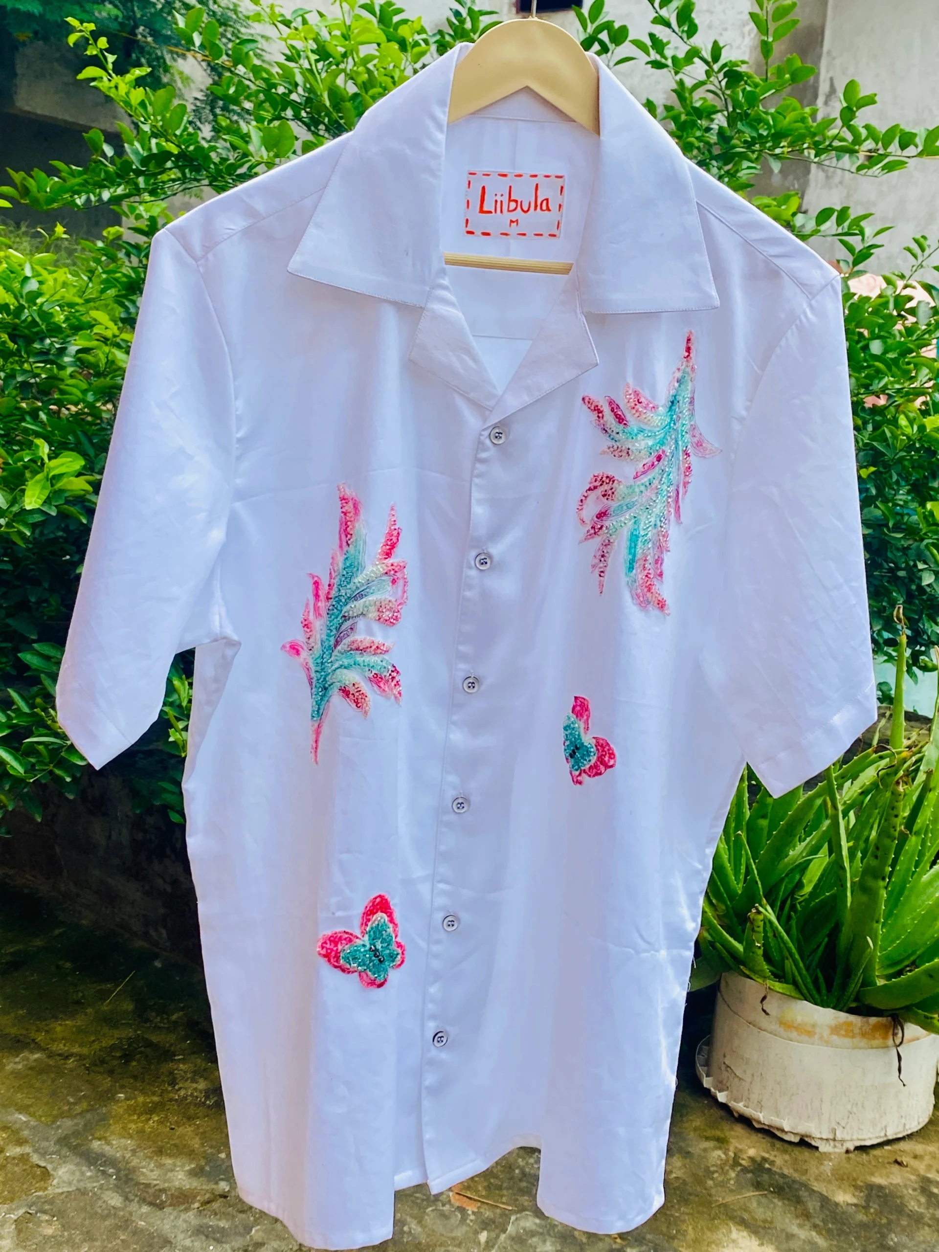 Full view of Le Butterfly Hand Embroidery Shirt, featuring white cotton fabric with vibrant hand embroidery, displayed outdoors on a hanger.
