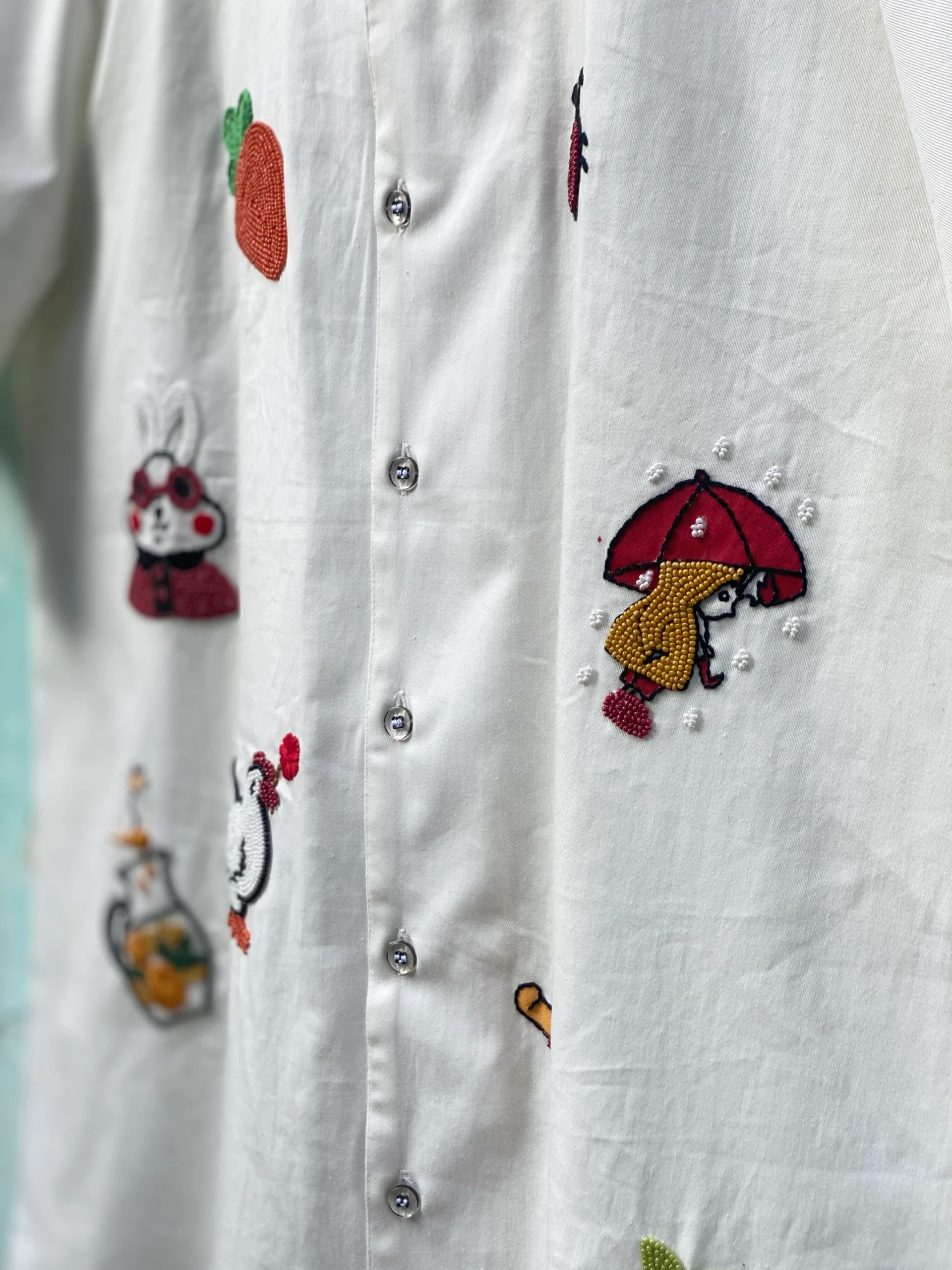 "Handmade cotton shirt with colorful hand embroidery, moti work, and fabric paint featuring motifs like bunny, umbrella girl, cherries, chicken, and carrot – limited edition Cuban collar shirt by Liibula"
