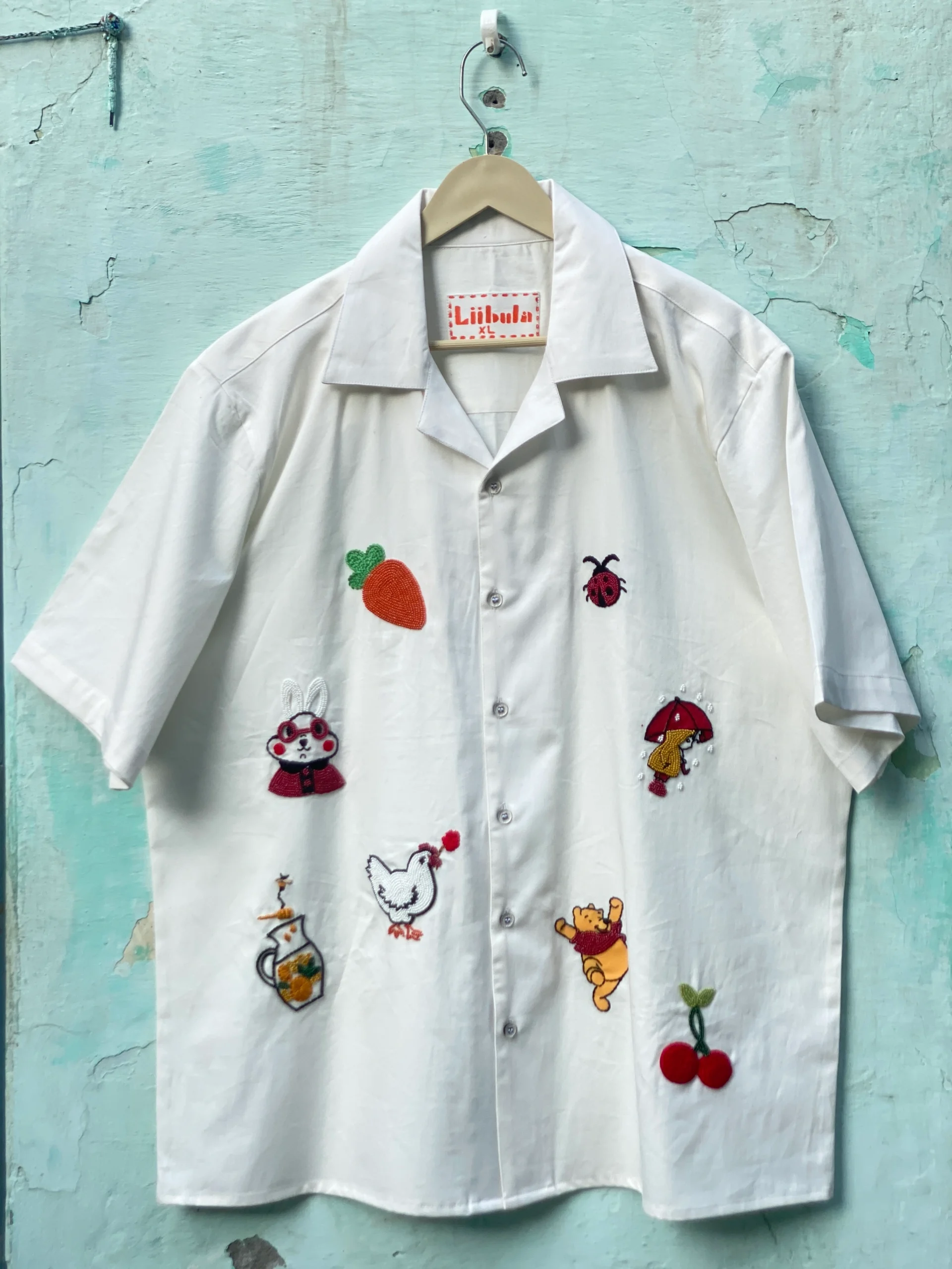 "Handmade cotton shirt with colorful hand embroidery, moti work, and fabric paint featuring motifs like bunny, umbrella girl, cherries, chicken, and carrot – limited edition Cuban collar shirt by Liibula"
