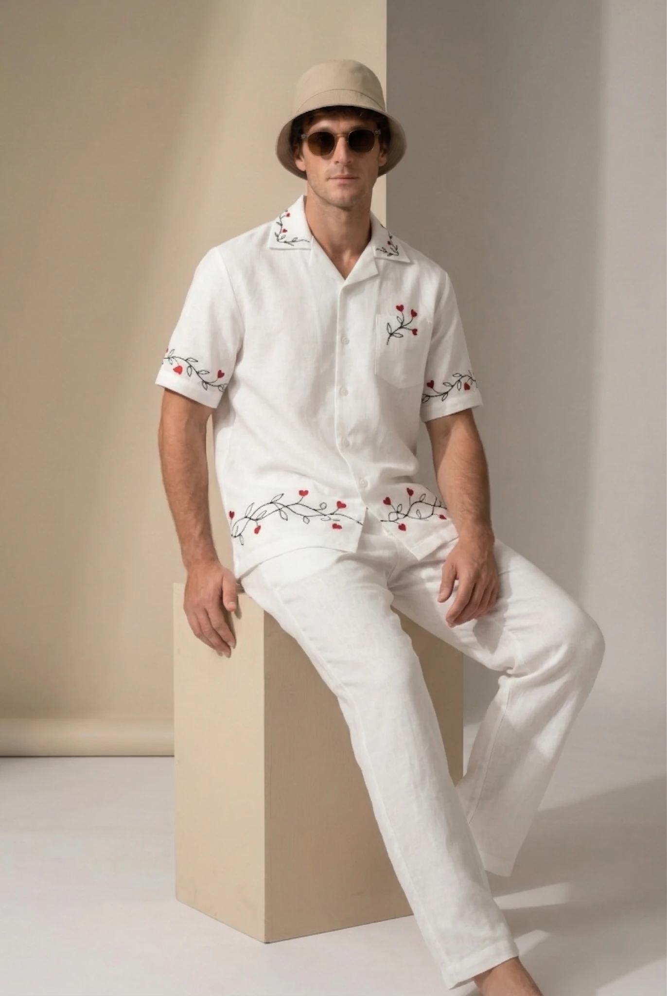 Men white embroidered linen shirt sitting pose in studio, red heart vine embroidery detail, luxury summer outfit with relaxed fit pants